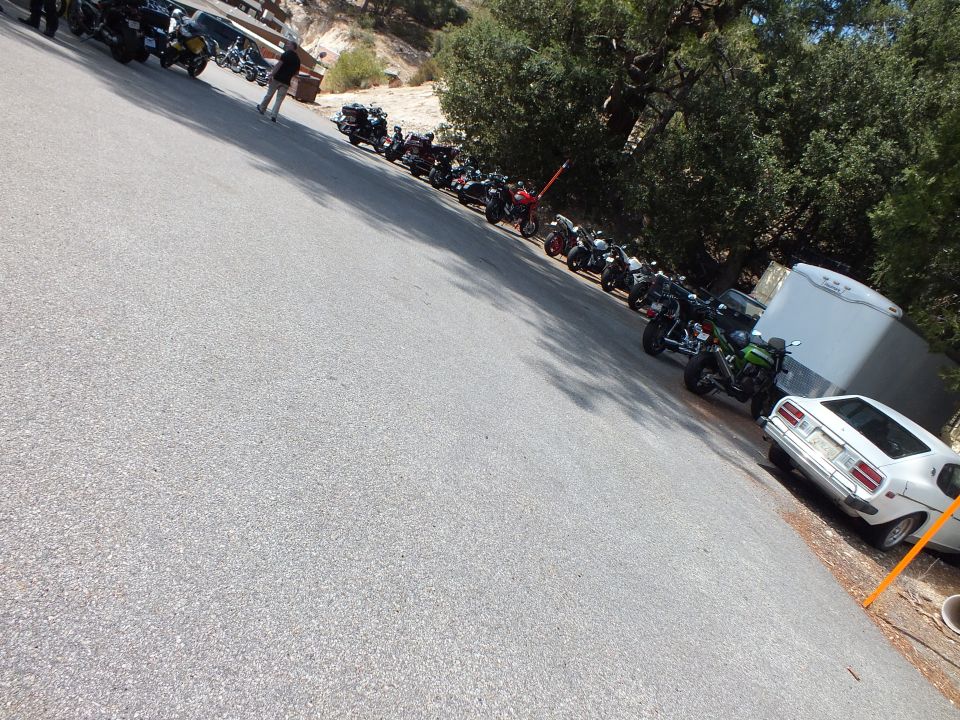 Line of parked motorcycles along a paved lot edge with white coupe and trailer