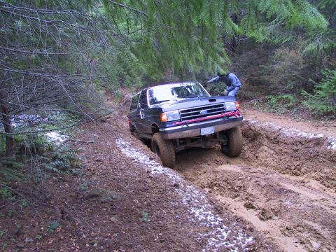 1991 Ford Bronco - A dark-colored Ford Bronco is stuck in a muddy rut on a wooded trail, with a person observing from the side.