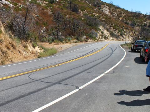 2003 Ford Crown Victoria - Long black tire marks sweep across a winding hillside highway, cars parked at the bend.
