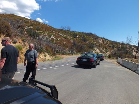 2003 Ford Crown Victoria - Onlookers stand by a pullout as a dark sedan rolls along the hillside road.