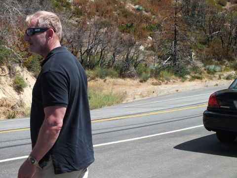 2003 Ford Crown Victoria - Profile of a man in sunglasses by a road, rear of a dark sedan visible at right.