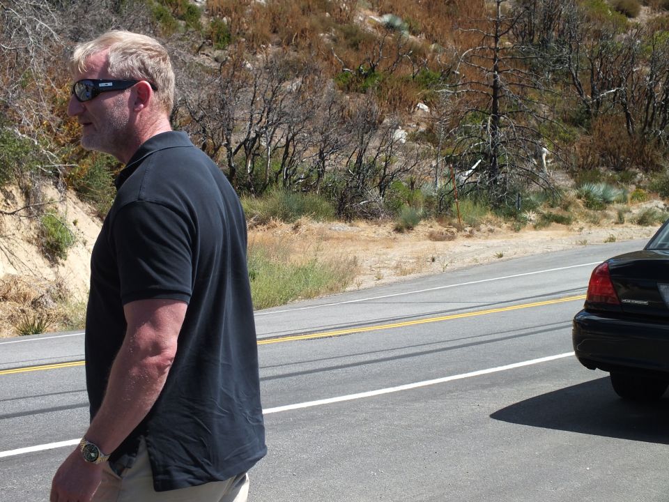 Man in black polo and sunglasses standing beside a black sedan on a roadside