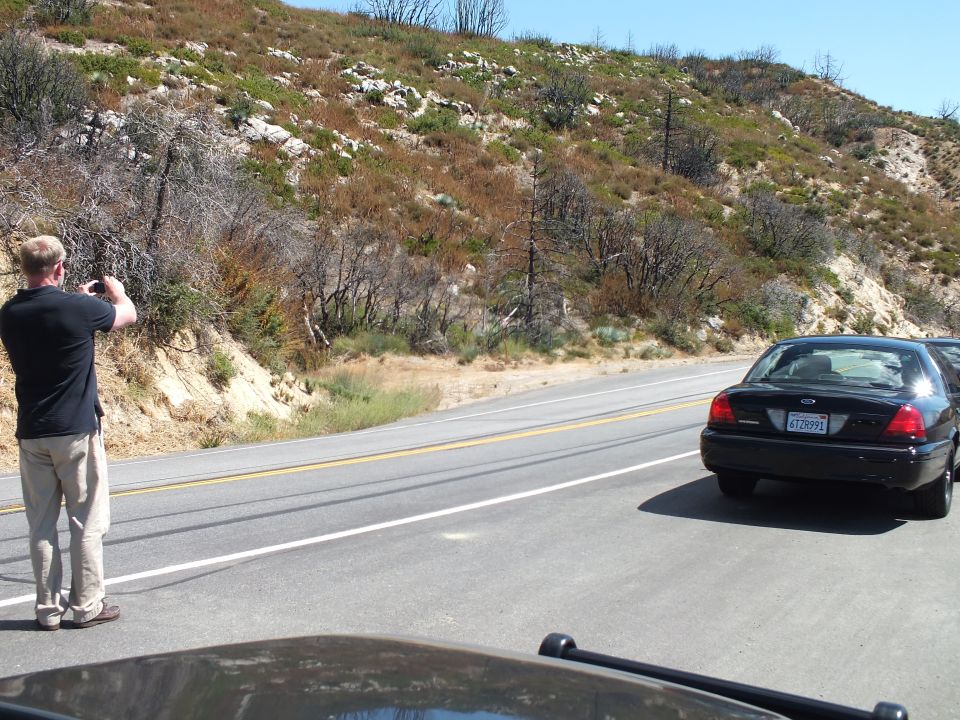 Man photographing a passing black Ford Crown Victoria on a hillside highway