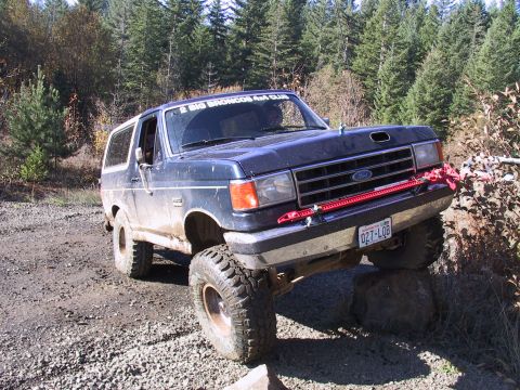 1991 Ford Bronco - A dark blue Ford Bronco navigates an off-road obstacle course with large tires and a red jack mounted on the front.