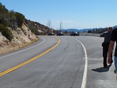 2003 Ford Crown Victoria - Pair heads down a curving road toward three dark sedans staged near a concrete barrier.