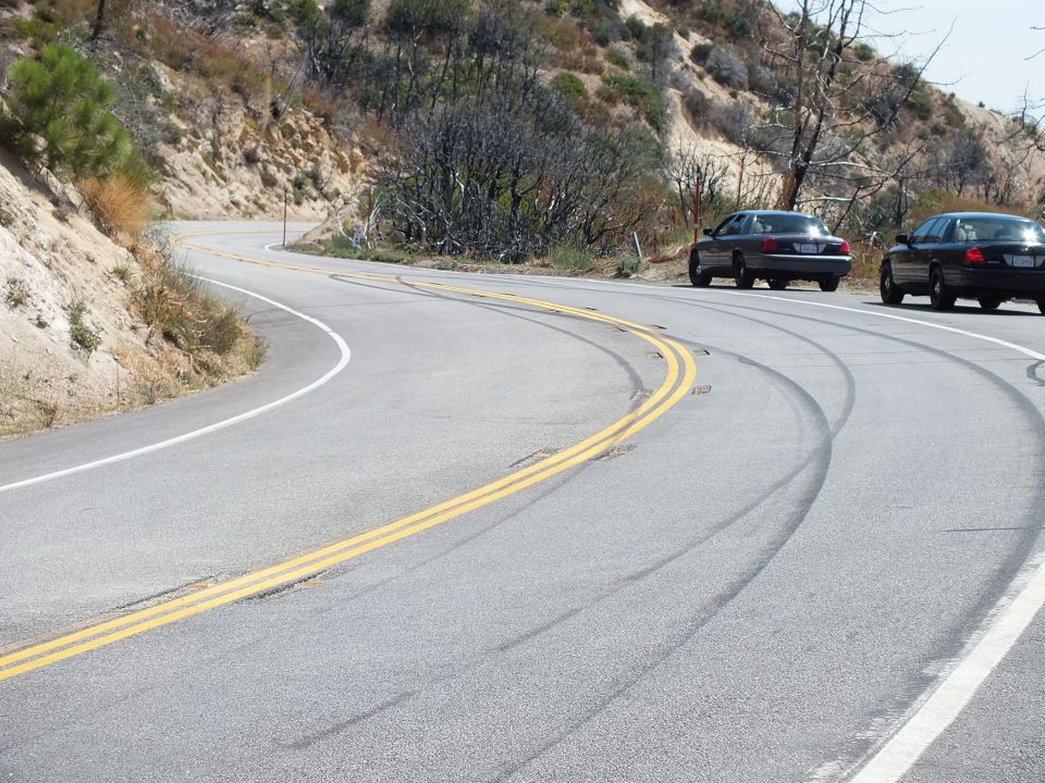 Heavy tire skid marks curving across a mountain highway with black sedans parked