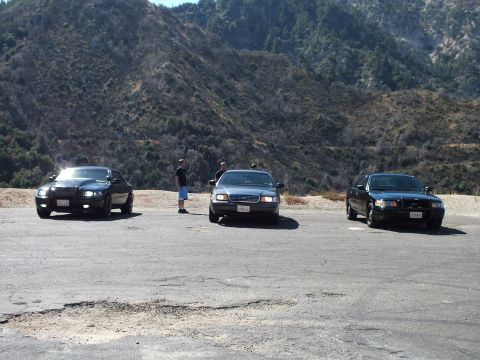 2003 Ford Crown Victoria - Trio of dark sedans spread across a dirt pullout under brush-covered slopes.