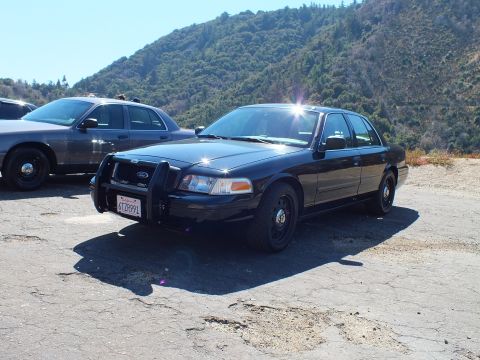 2003 Ford Crown Victoria - Black full-size Ford sedan with front push bar parked beside a darker sedan on cracked pavement