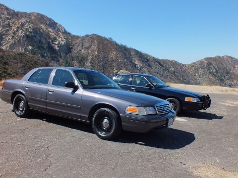 2003 Ford Crown Victoria - Two full-size Ford sedans, one gray and one black with push bar, on a hillside pullout