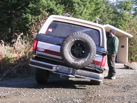 1991 Ford Bronco - Rear view of a white Ford Bronco full-size SUV with a red and silver tailgate stripe, parked at an angle on a gravel forest-road pullout wi…