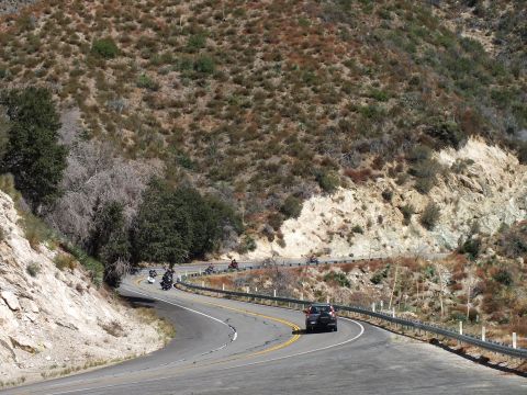 2003 Ford Crown Victoria - Riders and a vehicle navigating a curving two-lane road cut into a chaparral hillside