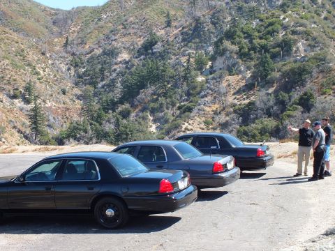 2003 Ford Crown Victoria - Three men stand beside a line of black Panther-platform sedans at a canyon overlook.