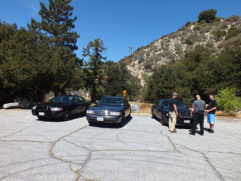 2003 Ford Crown Victoria - Three Panther-platform sedans sit in a canyon parking lot as men gather beside the rightmost car.