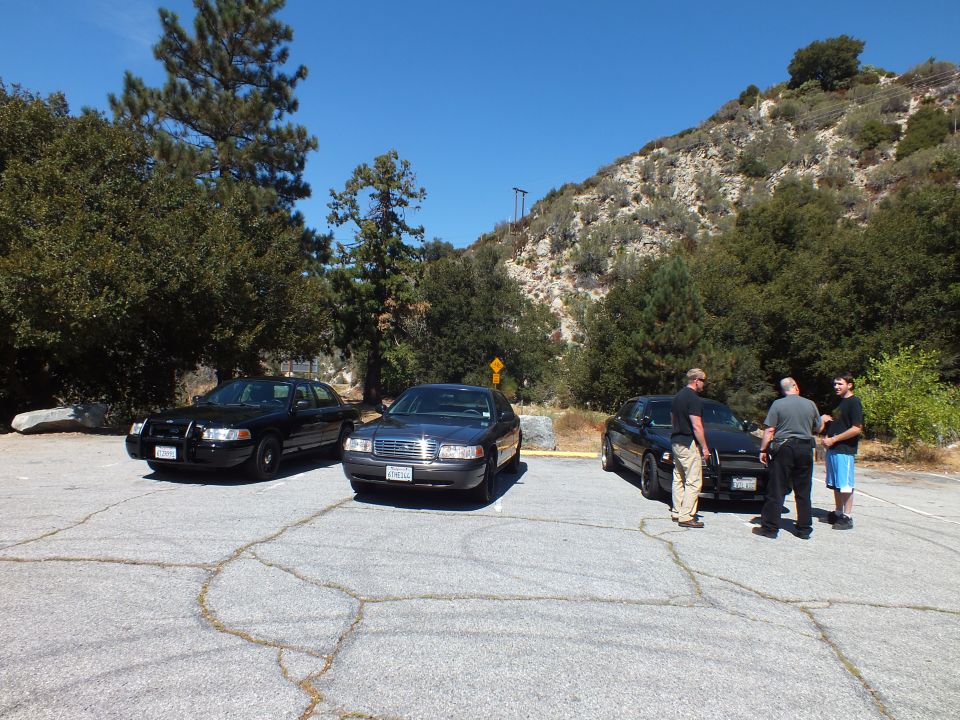 Three dark sedans parked in cracked asphalt mountain lot with three men by one car