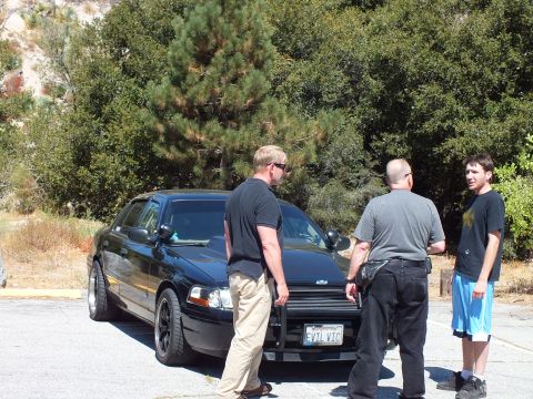 2003 Ford Crown Victoria - Men stand beside a blacked-out Ford sedan whose California plate reads EVIL VIC, grille guard fitted.
