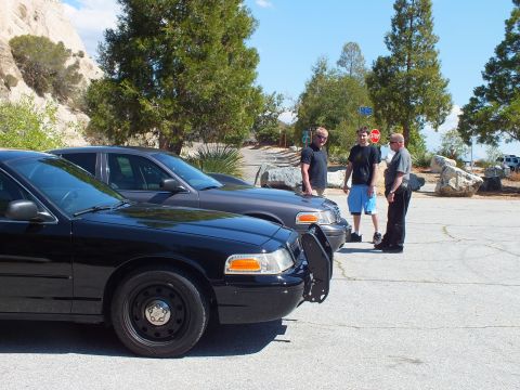 2003 Ford Crown Victoria - Front corners of two blacked-out Panther sedans face the camera as people gather by the rocks.