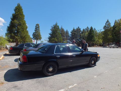2003 Ford Crown Victoria - Rear quarter of a blacked-out full-size Ford sedan on cracked asphalt with people in the distance.