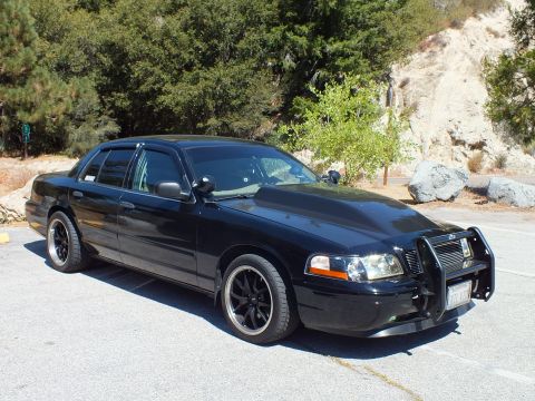 2003 Ford Crown Victoria - lowered blacked-out Ford sedan with grille guard sits on asphalt against a rocky canyon slope.