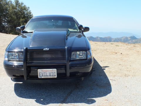 2003 Ford Crown Victoria - blacked-out Crown Victoria with grille guard parks on a summit, plate EVTL VTC, hazy ridges beyond.
