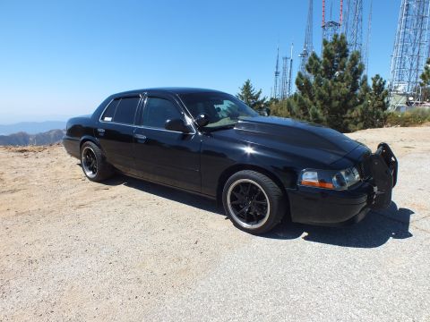 2003 Ford Crown Victoria - blacked-out Ford sedan with grille guard sits atop a peak with broadcast masts behind it.