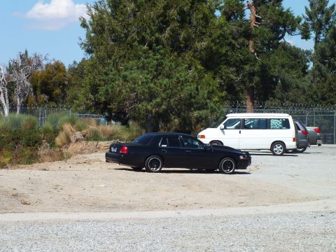 2003 Ford Crown Victoria - Lowered black sedan with black wheels sits beside a white van on a dirt lot