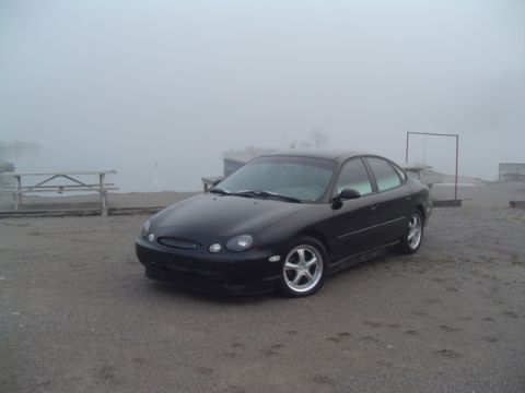 1999 Ford Taurus - A black sedan sits parked near a body of water shrouded in thick fog.