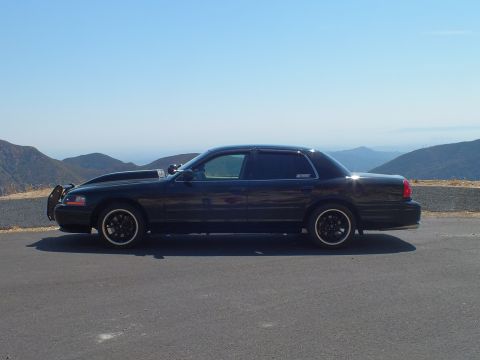 2003 Ford Crown Victoria - Side view of a lowered black Mercury sedan parked at a hilltop turnout, distant ranges behind.