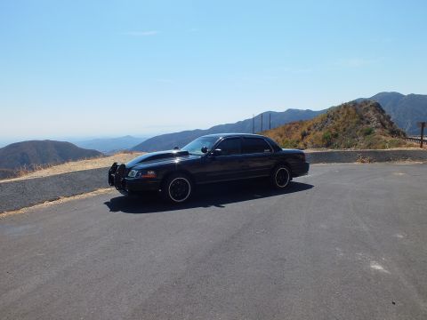 2003 Ford Crown Victoria - Black Mercury sedan with front push bar at a paved hilltop pullout, ranges in distance.