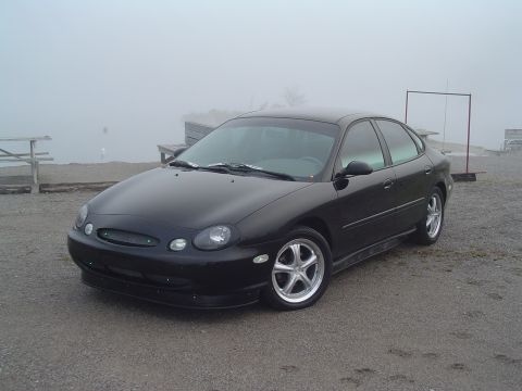 1999 Ford Taurus - This black 1999 Ford Taurus is seen from a three-quarter front view on a gravel lot with a foggy background.