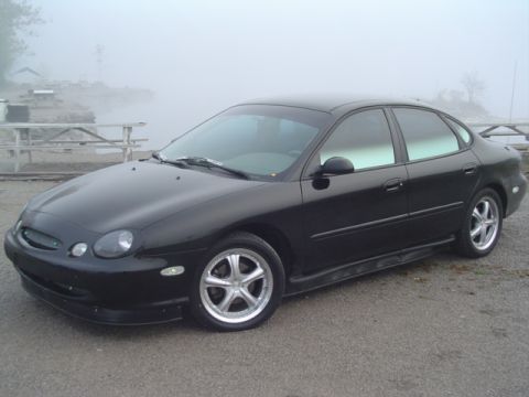 1999 Ford Taurus - A black 1999 Ford Taurus sedan is parked on a gravel lot near a body of water on a foggy day.