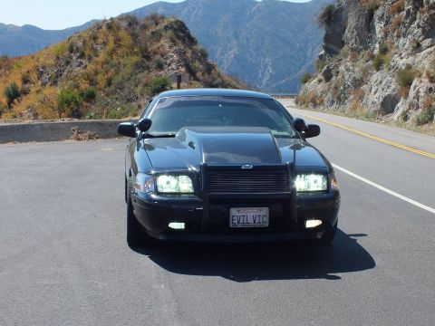 2003 Ford Crown Victoria - Head-on shot of a blacked-out Mercury with hood scoop, lights on, reading EVIL VIC plate.