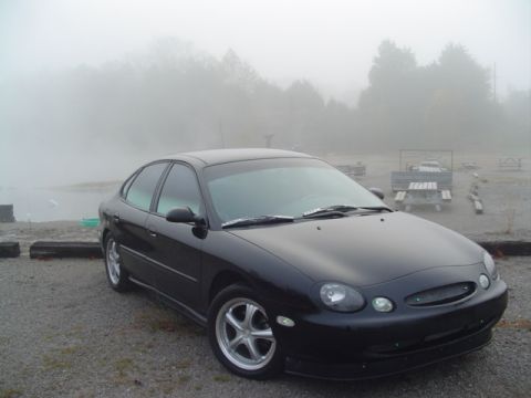 1999 Ford Taurus - A black sedan is parked in a lot with picnic tables visible in the background, obscured by fog.