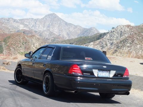 2003 Ford Crown Victoria - Rear three-quarter of a black Mercury sedan showing taillights and EVIL VIC plate, mountains beyond.