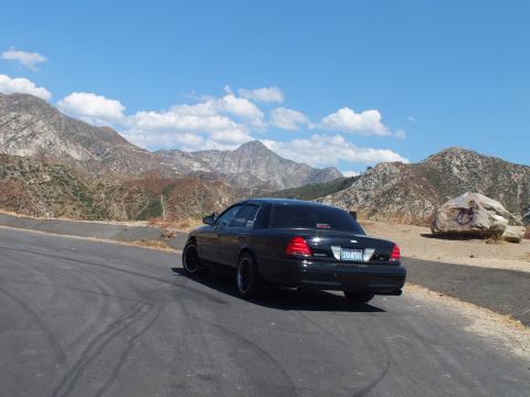 2003 Ford Crown Victoria - Rear view of a blacked-out Mercury sedan parked at a hilltop pullout, rugged peaks behind.