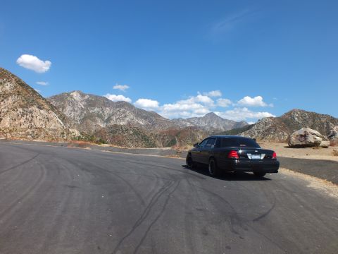 2003 Ford Crown Victoria - Distant black Mercury sedan at a paved turnout framed by mountains and scattered clouds.