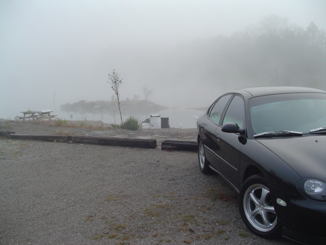 Black 1999 Ford Taurus parked on gravel near a lake with picnic tables in the fog