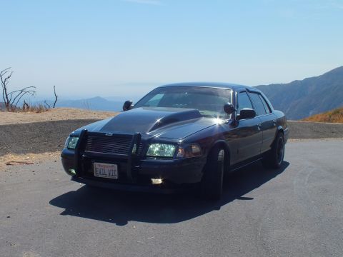 2003 Ford Crown Victoria - Front-quarter view of a blacked-out Crown Vic showing push bar and lit headlights on a mountain road.