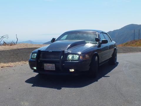 2003 Ford Crown Victoria - Crown Vic angled front view with grille guard and headlights on, valley backdrop.