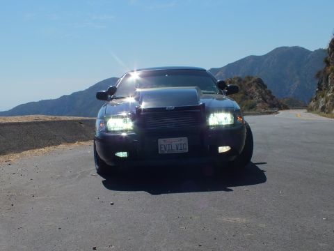 2003 Ford Crown Victoria - Direct front shot of a blacked-out Crown Vic, sun glare on hood, mountain road behind.