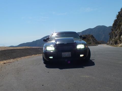 2003 Ford Crown Victoria - Front view of a murdered-out Crown Vic, headlights glowing against blue sky and ridge.