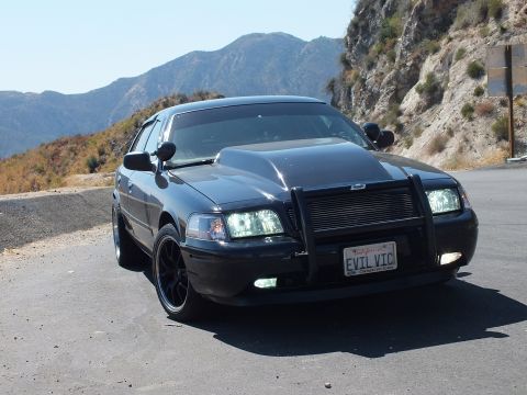 2003 Ford Crown Victoria - Blacked-out Crown Vic angled toward camera, grille guard and fog lights visible.
