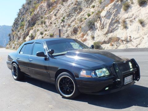 2003 Ford Crown Victoria - Crown Vic front-quarter showing long hood and grille guard at a rocky roadside.