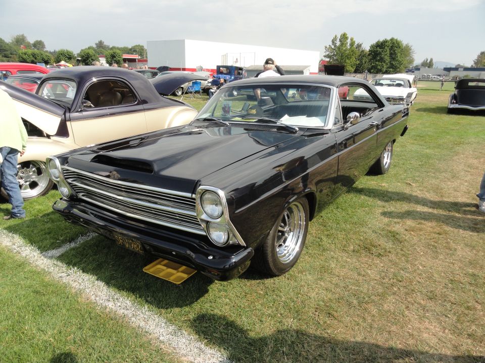 Black Ford Fairlane hardtop with hood scoop and polished mag wheels at a grass car show