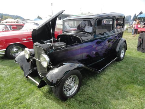 1964 Ford Fairlane - A black hot rod sedan with flame decals is displayed at a car show next to a red car.