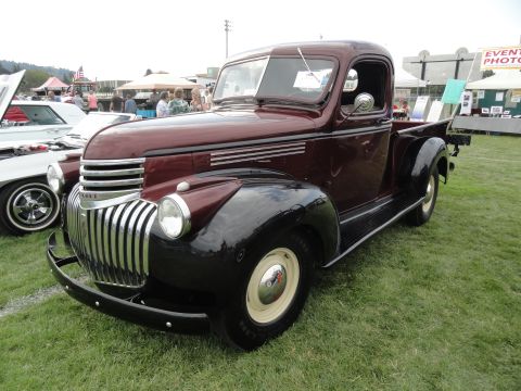 1964 Ford Fairlane - A maroon and black pickup truck is parked on a grassy field at an outdoor car show.