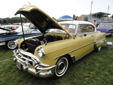 1964 Ford Fairlane - A yellow 1954 Chevrolet Bel Air coupe is displayed with its hood open at an outdoor car show.