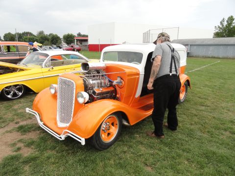 1964 Ford Fairlane - A man inspects an orange and white hot rod at a car show, with another classic car visible to the left.