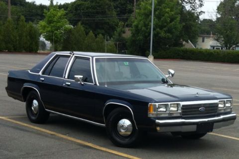 1991 Ford LTD Crown Victoria - A dark blue sedan with chrome accents is parked in an empty lot under a cloudy sky.