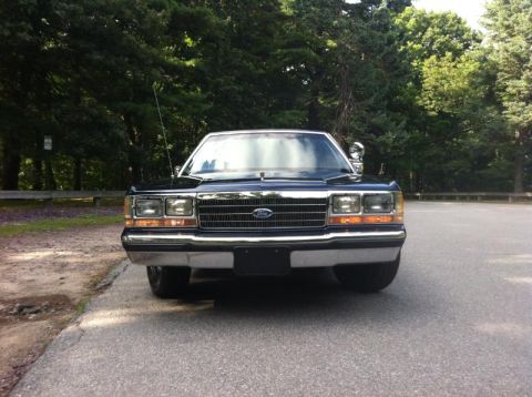1991 Ford LTD Crown Victoria - Front view of a black Ford Crown Victoria parked on asphalt with trees in the background.
