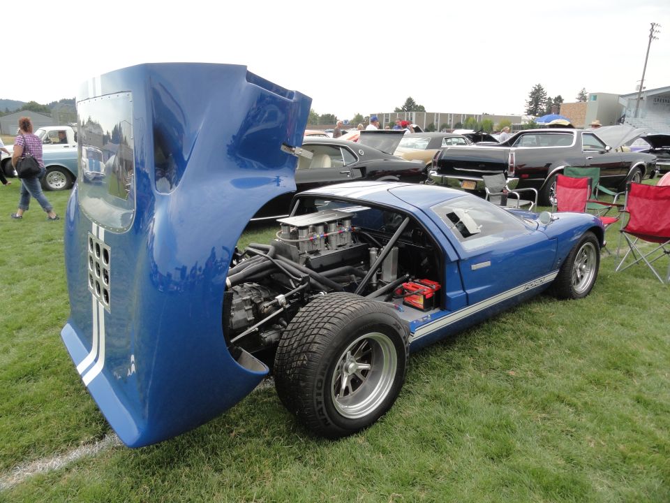 Blue race car with hood open, showing engine and battery. White stripes on hood and side. Other cars and chairs in background